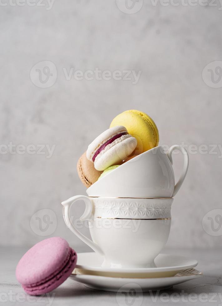 Stack of white coffee cups filled with macaroons on the edge of the table, white brick wall background photo