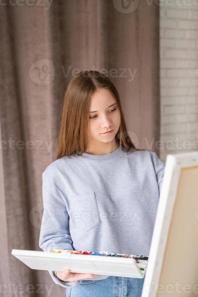 Young thoughtful female artist painting on the canvas in the studio holding a palette with watercolors photo