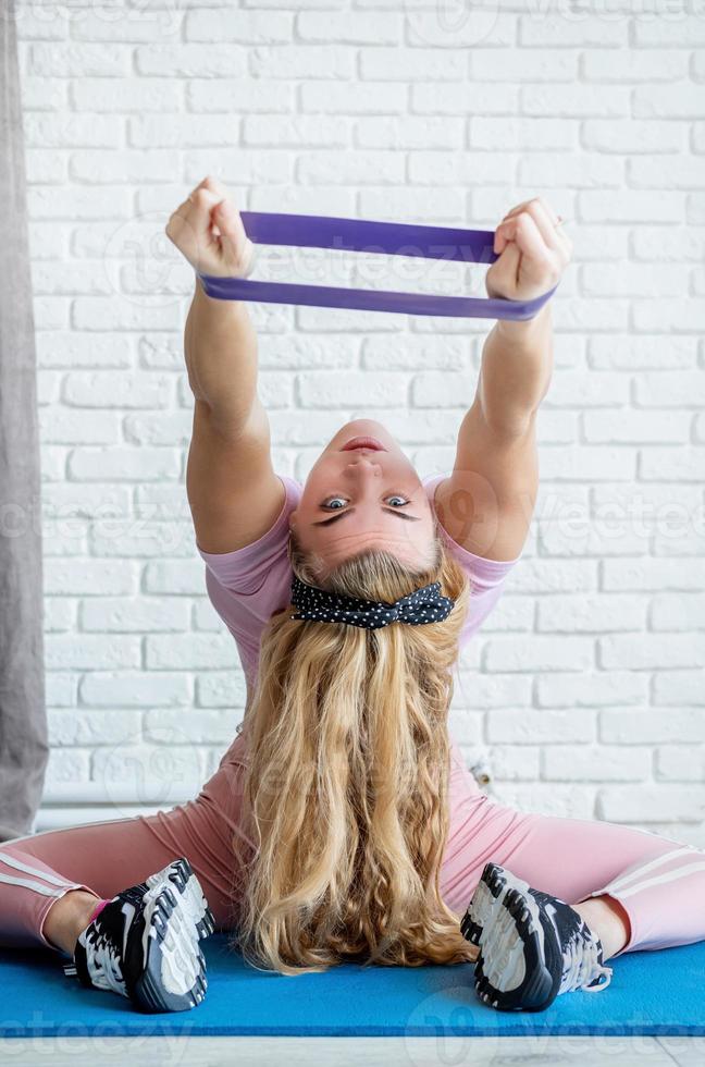 Mujer atlética haciendo ejercicios con una banda de resistencia sobre una colchoneta de fitness en casa con fondo de pared de ladrillo blanco foto