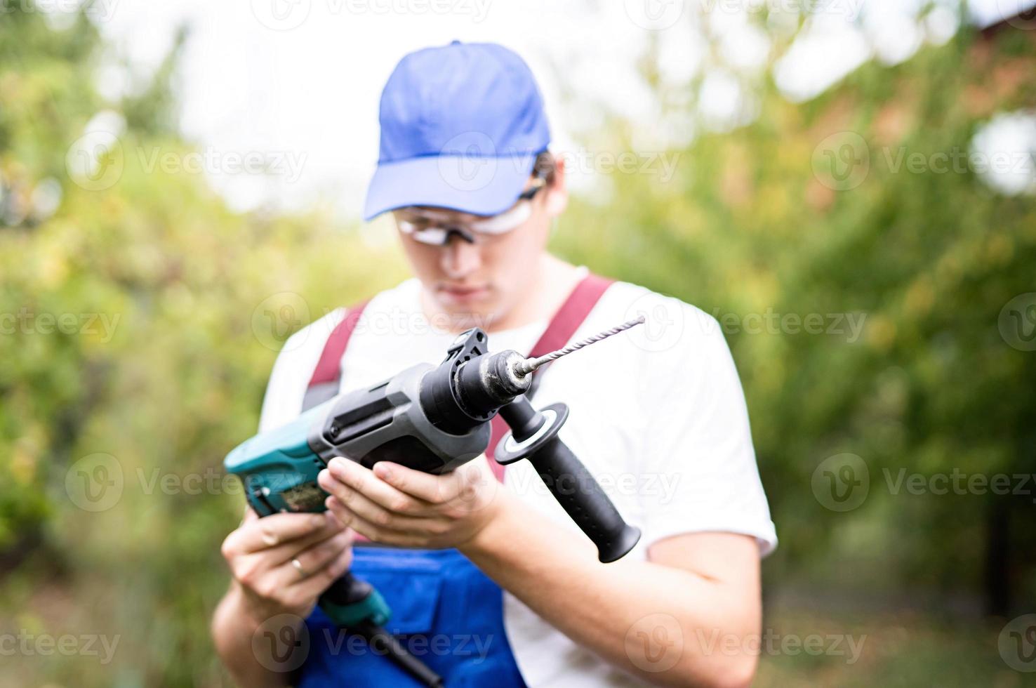 trabajador de la construcción en gafas de protección y uniforme mirando el perforador. hombre con taladro foto