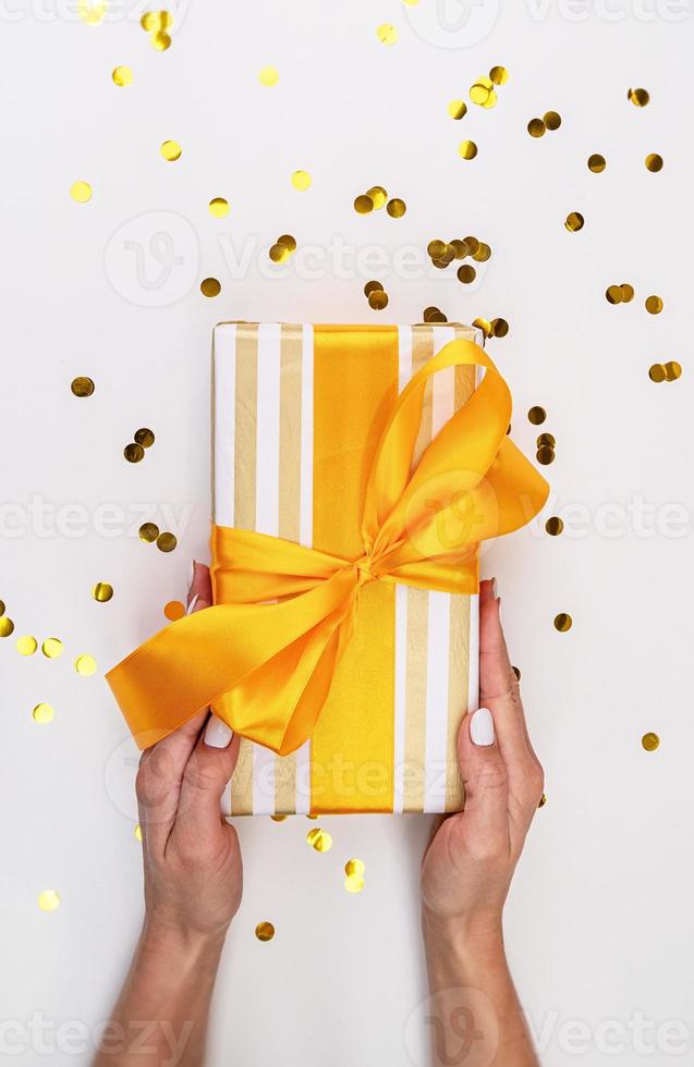 Woman holding a gift box wrapped in white and gold paper with confetti scattered around photo