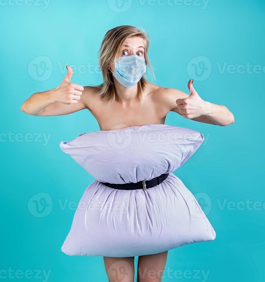Blond woman in pillowdress wearing a mask showing thumbs up isolated on blue background photo