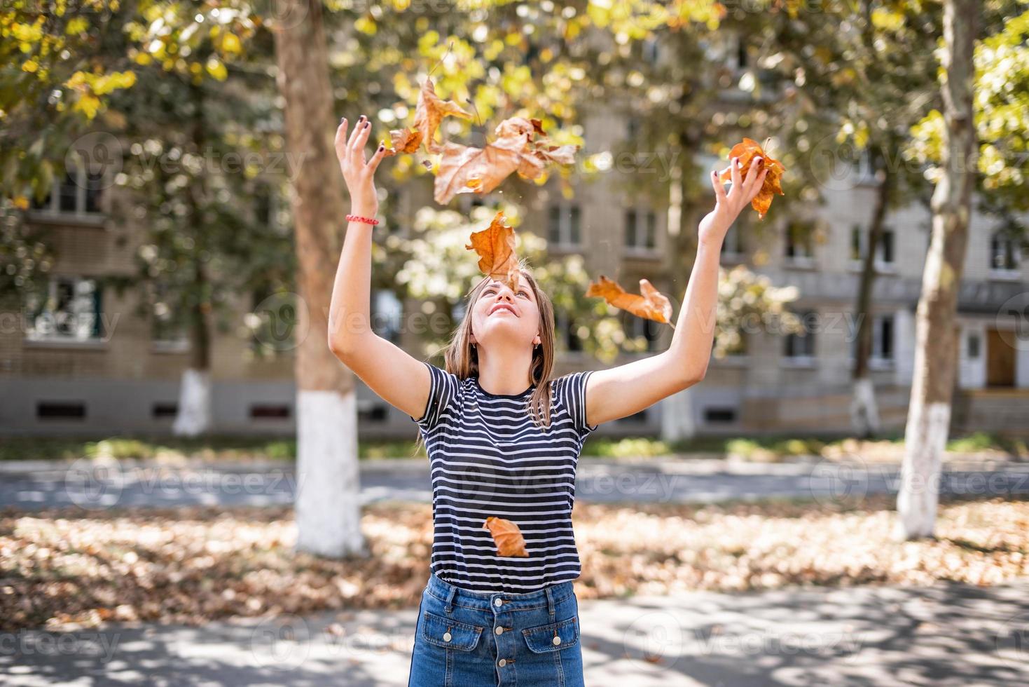 Bastante joven mujer caucásica arrojando hojas de otoño en el fondo del parque urbano foto