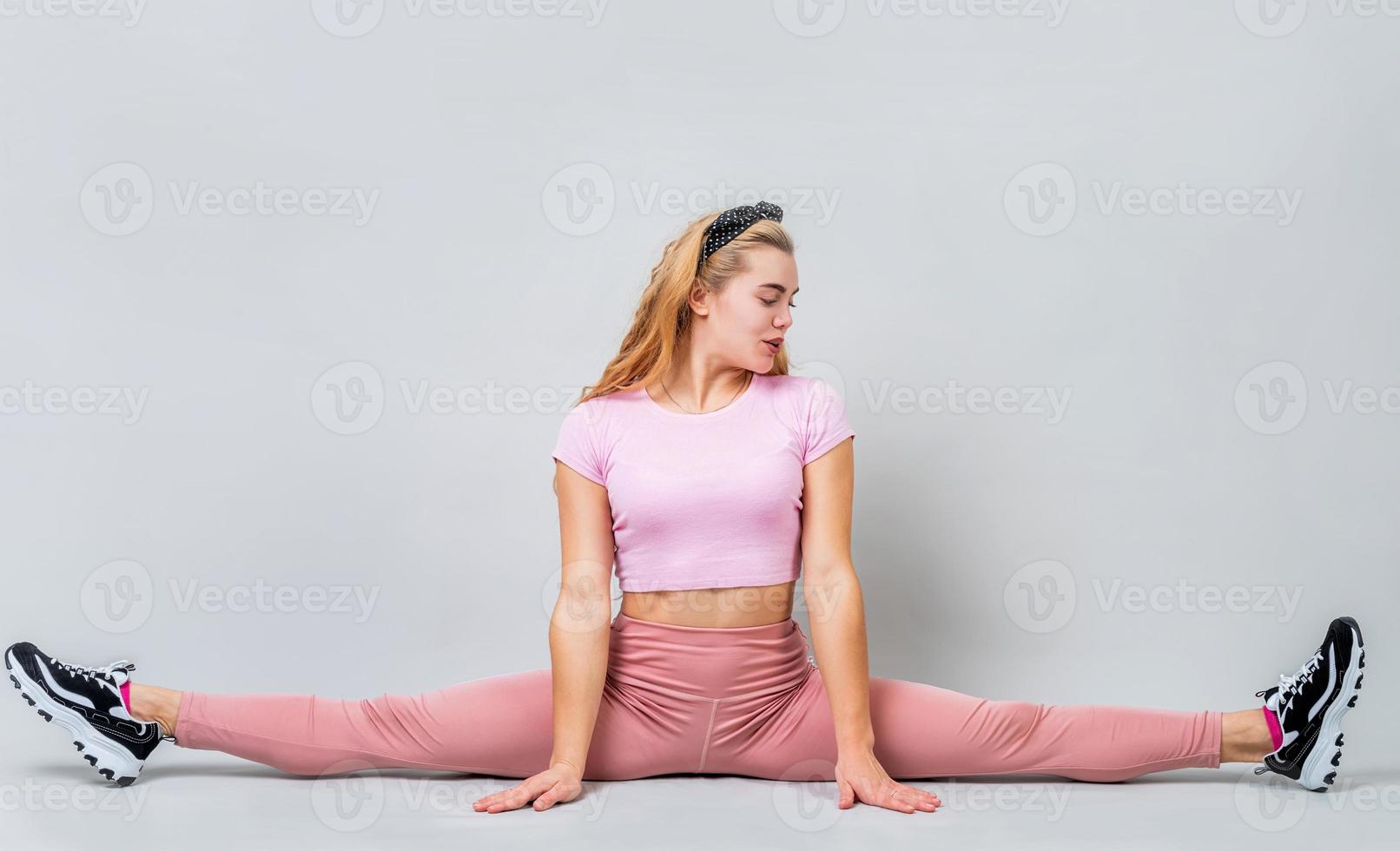 Mujer acróbata vistiendo ropa deportiva rosa que se extiende en una pose de pierna dividida en el estudio aislado sobre fondo gris foto