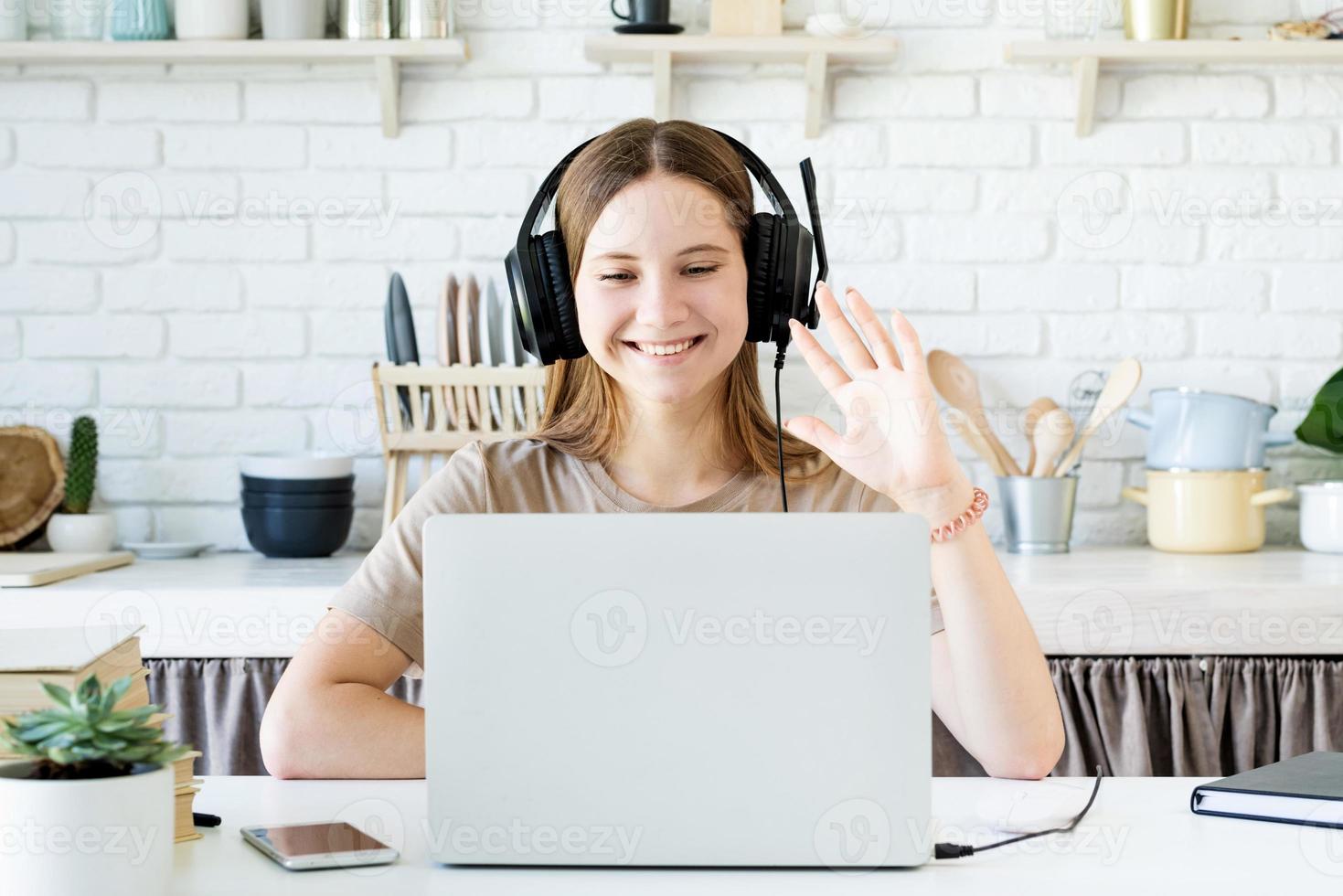 Sonriente jovencita sentada en el escritorio en la cocina aprendiendo usando su computadora portátil, saludando hola charlando con amigos foto