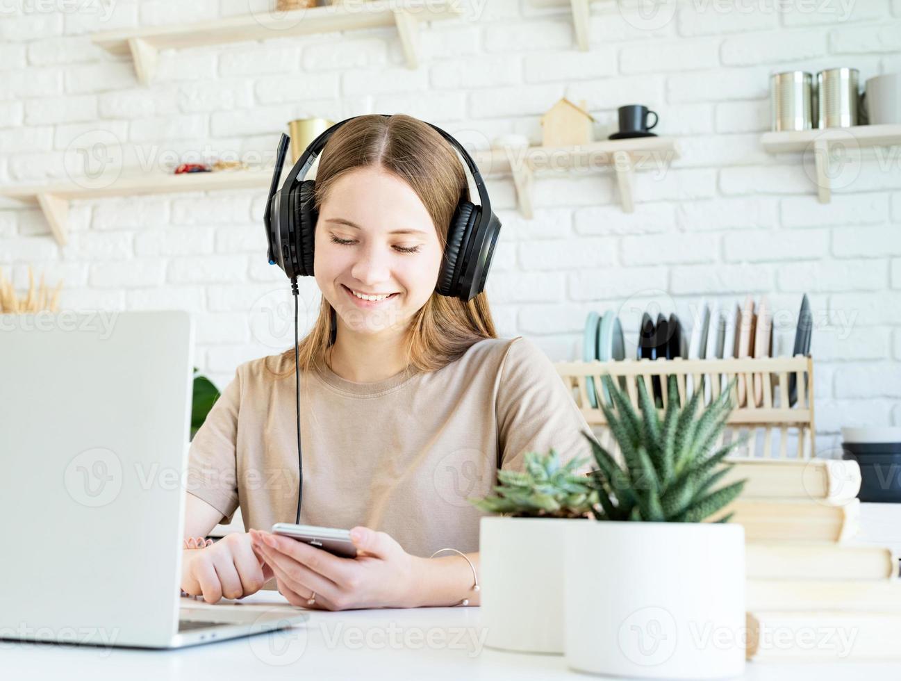 Sonriente jovencita en auriculares charlando con su smartphone mientras hace su homewotk foto