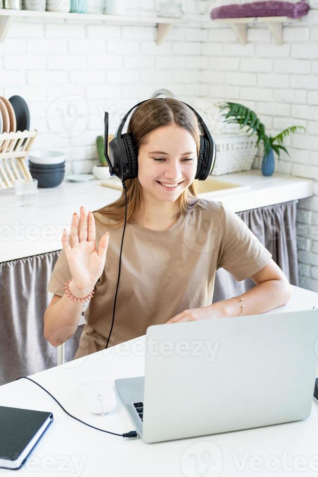 Sonriente jovencita sentada en el escritorio en la cocina aprendiendo usando su computadora portátil, saludando hola charlando con amigos foto