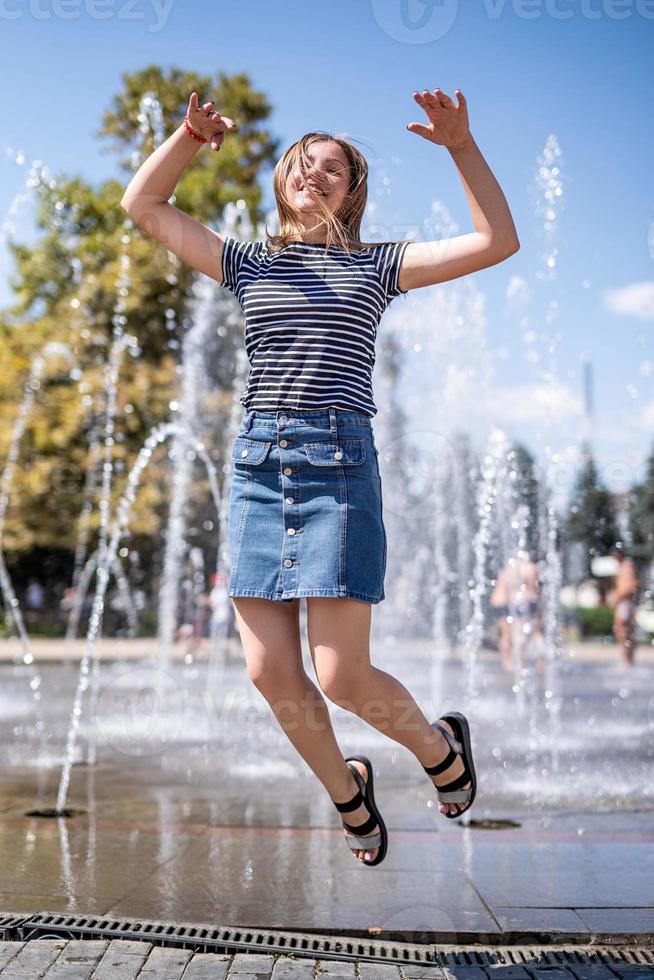 Joven mujer caucásica atractiva feliz saltando y riendo cerca de la fuente en el parque urbano foto