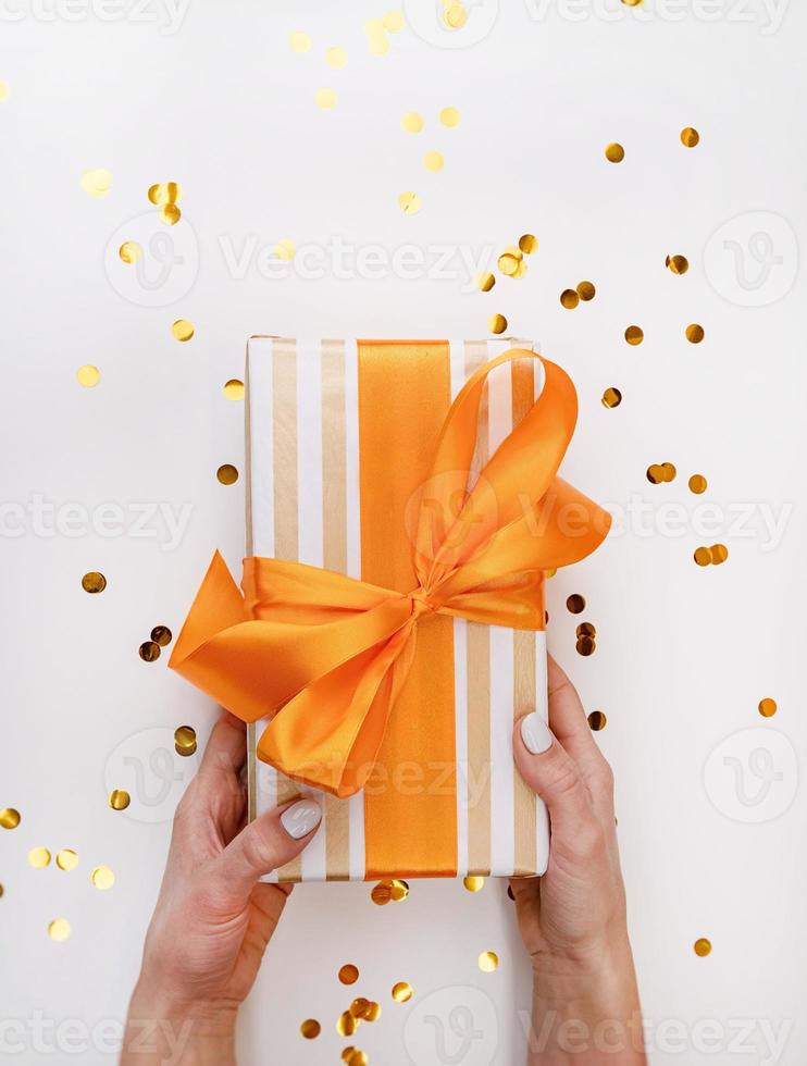 Female hands holding holiday gift wrapperd with white and gold paper with confetti scattered arounf flat lay photo