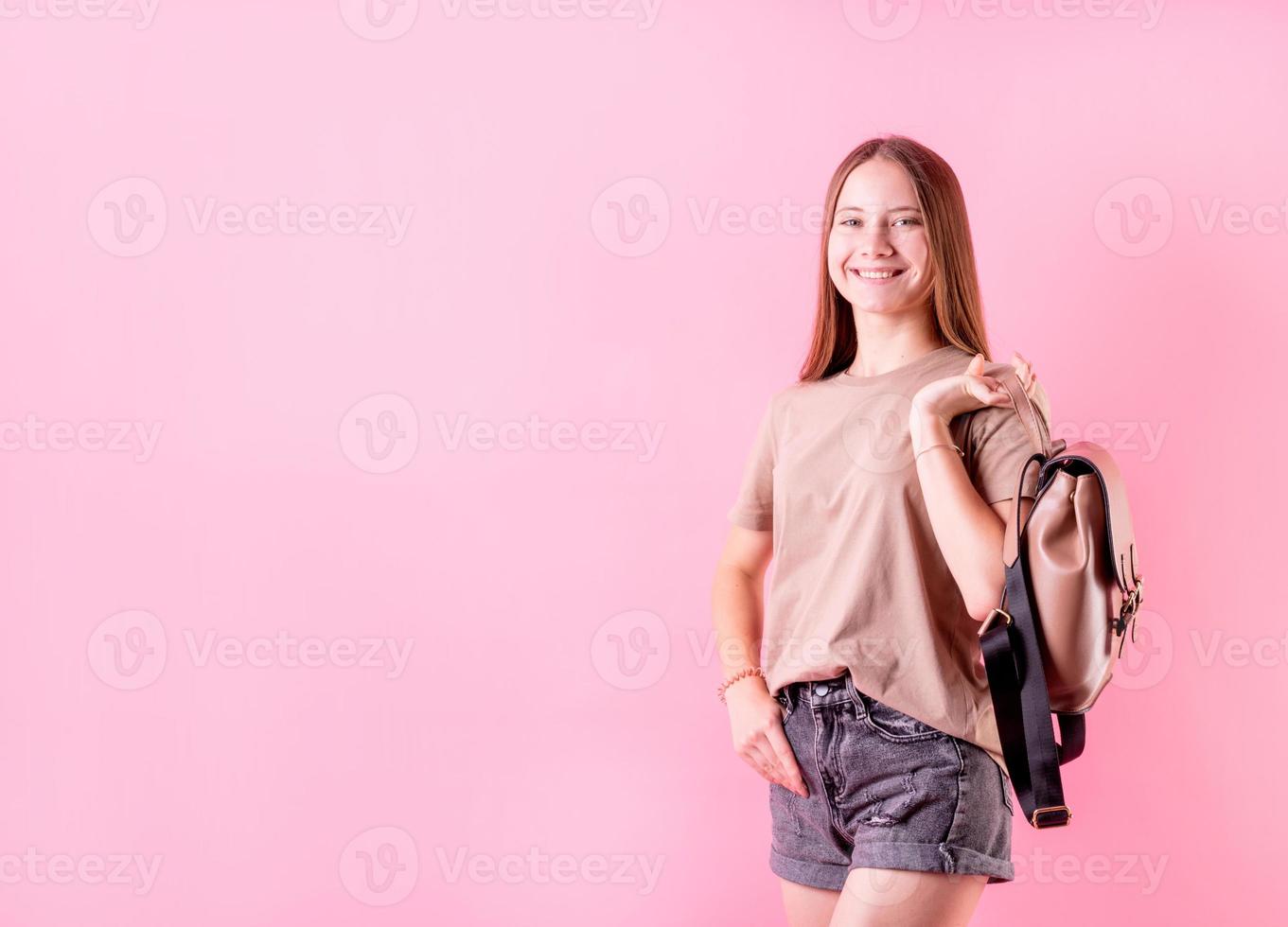 Feliz chica adolescente sosteniendo una mochila lista para la escuela aislada sobre fondo de color rosa foto