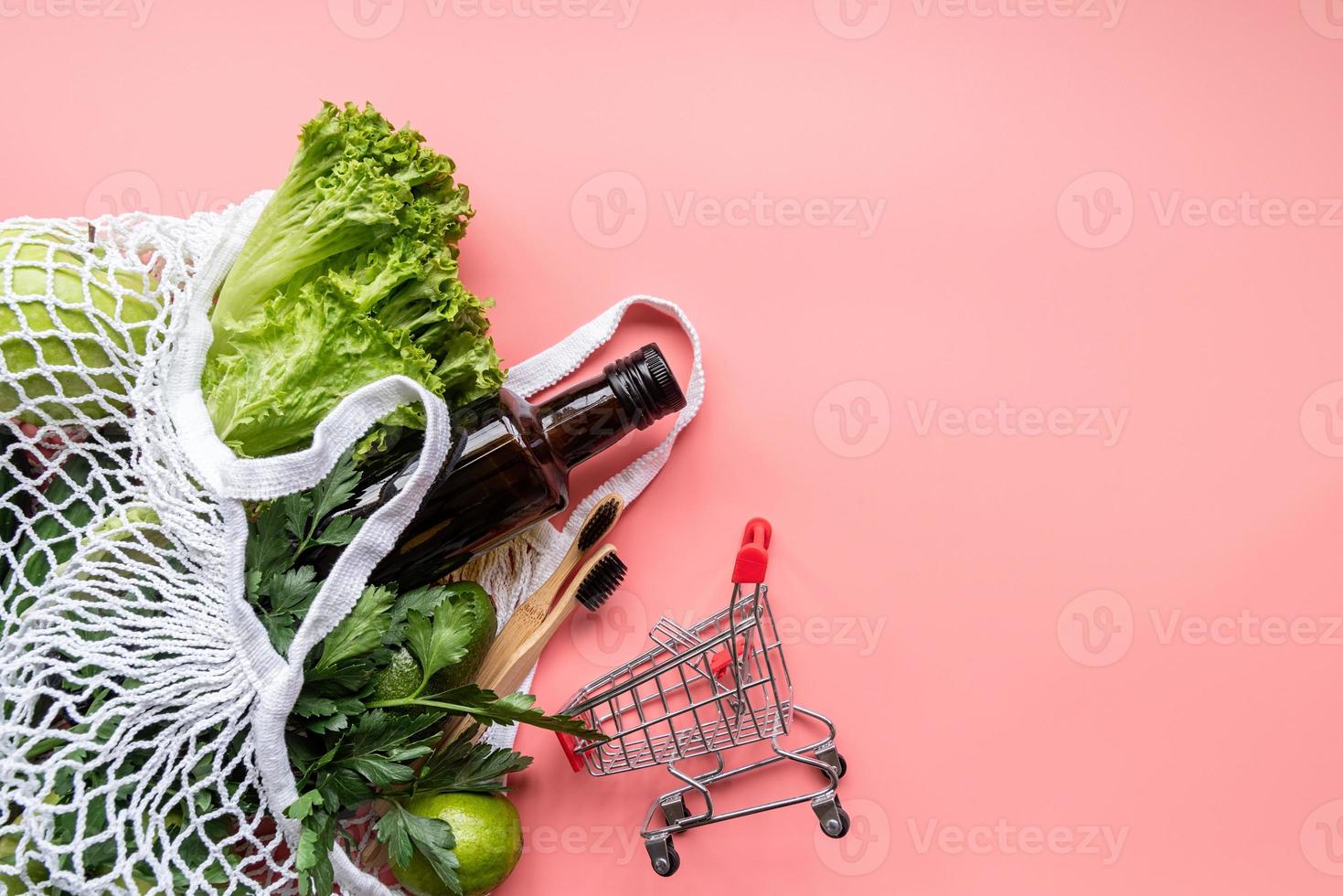 Bolsa de malla ecológica con verduras, cepillos de dientes de madera, guantes y vista superior de aceite de oliva sobre fondo rosa con espacio de copia foto