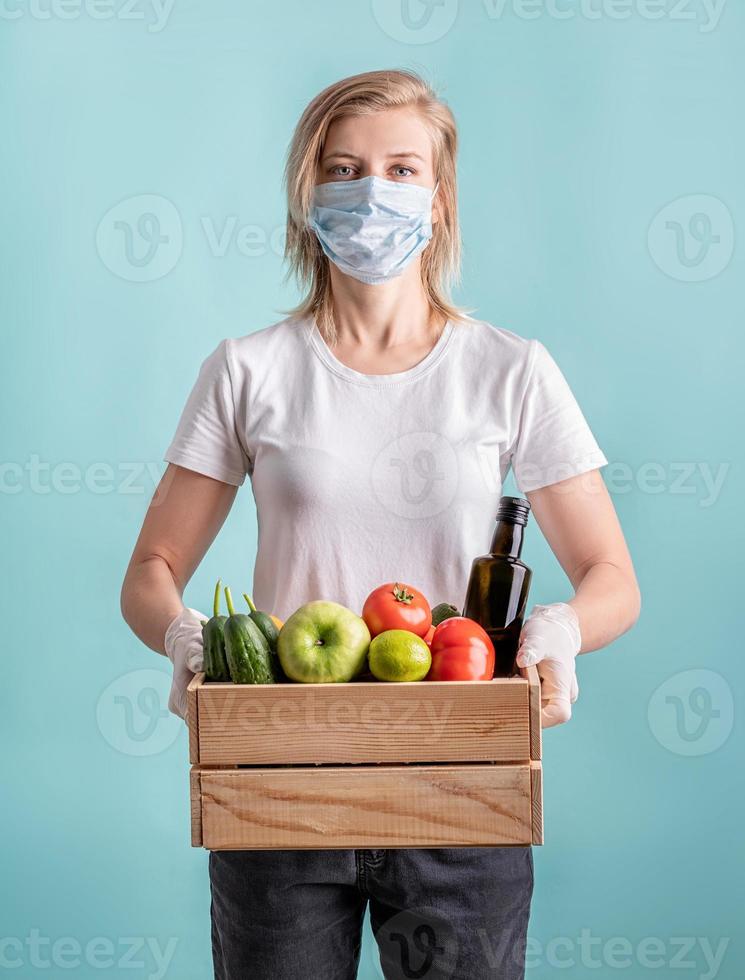 Blond woman in a mask and gloves holding a wooden box full of vegetables photo