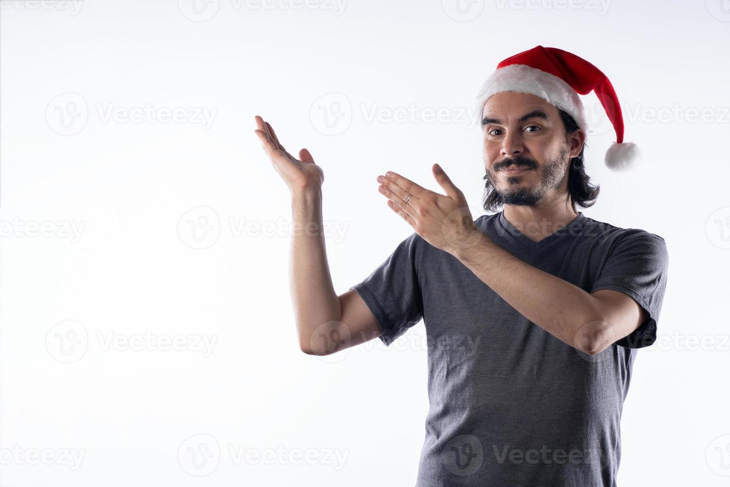 joven latino con sombrero de santa claus, con una sonrisa mostrando sus manos a los lados. Fondo blanco foto