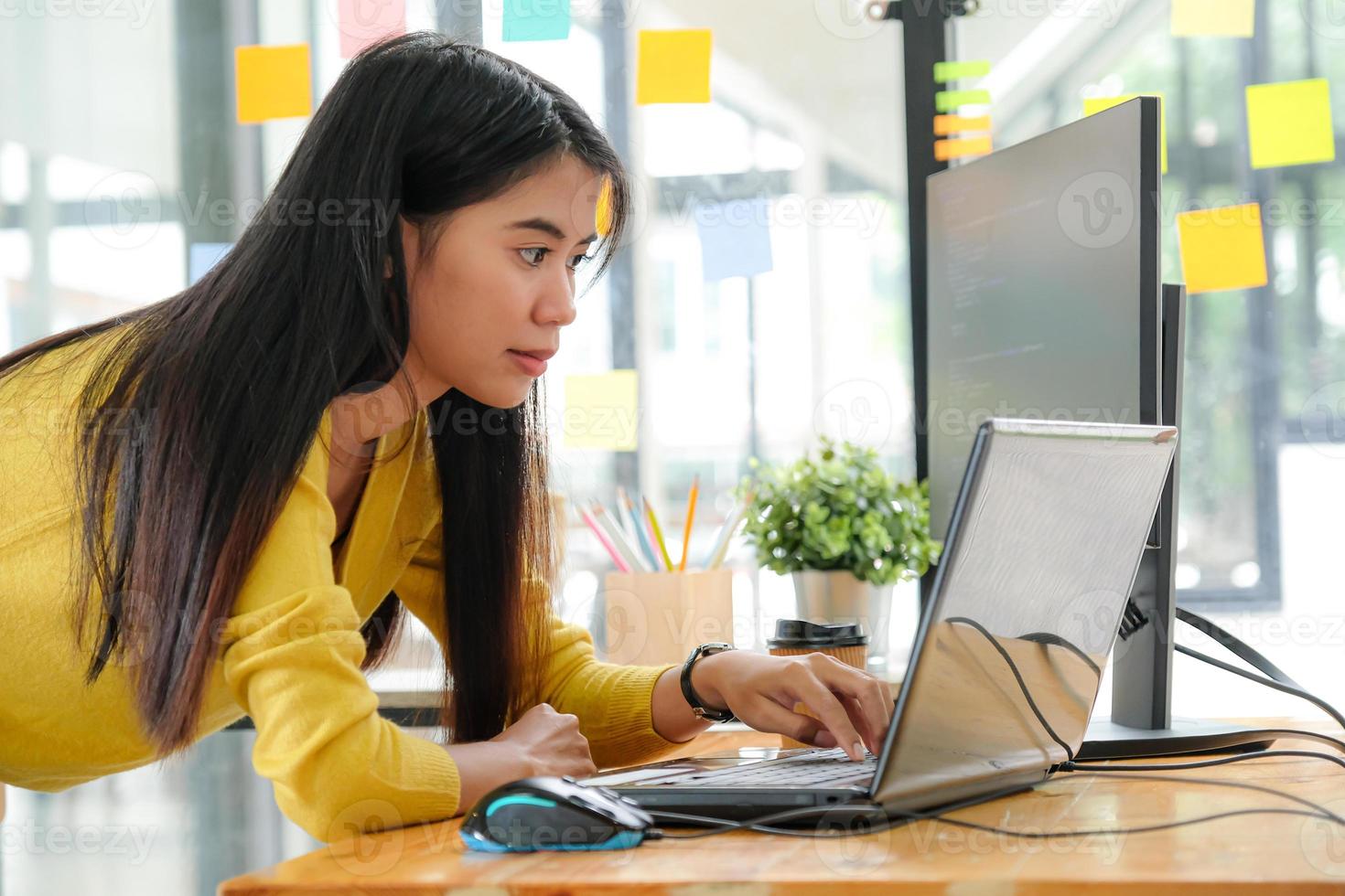 joven programadora asiática con una camisa amarilla se detiene para usar su computadora portátil y su PC en la oficina. foto