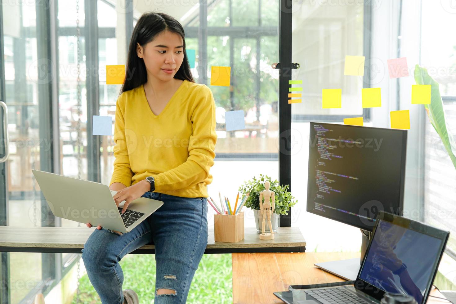 programadora asiática para camisa amarilla se sienta en el estante y coloca la computadora portátil en la pierna. Ella miró la pantalla de la computadora en la mesa y reflexionó. foto