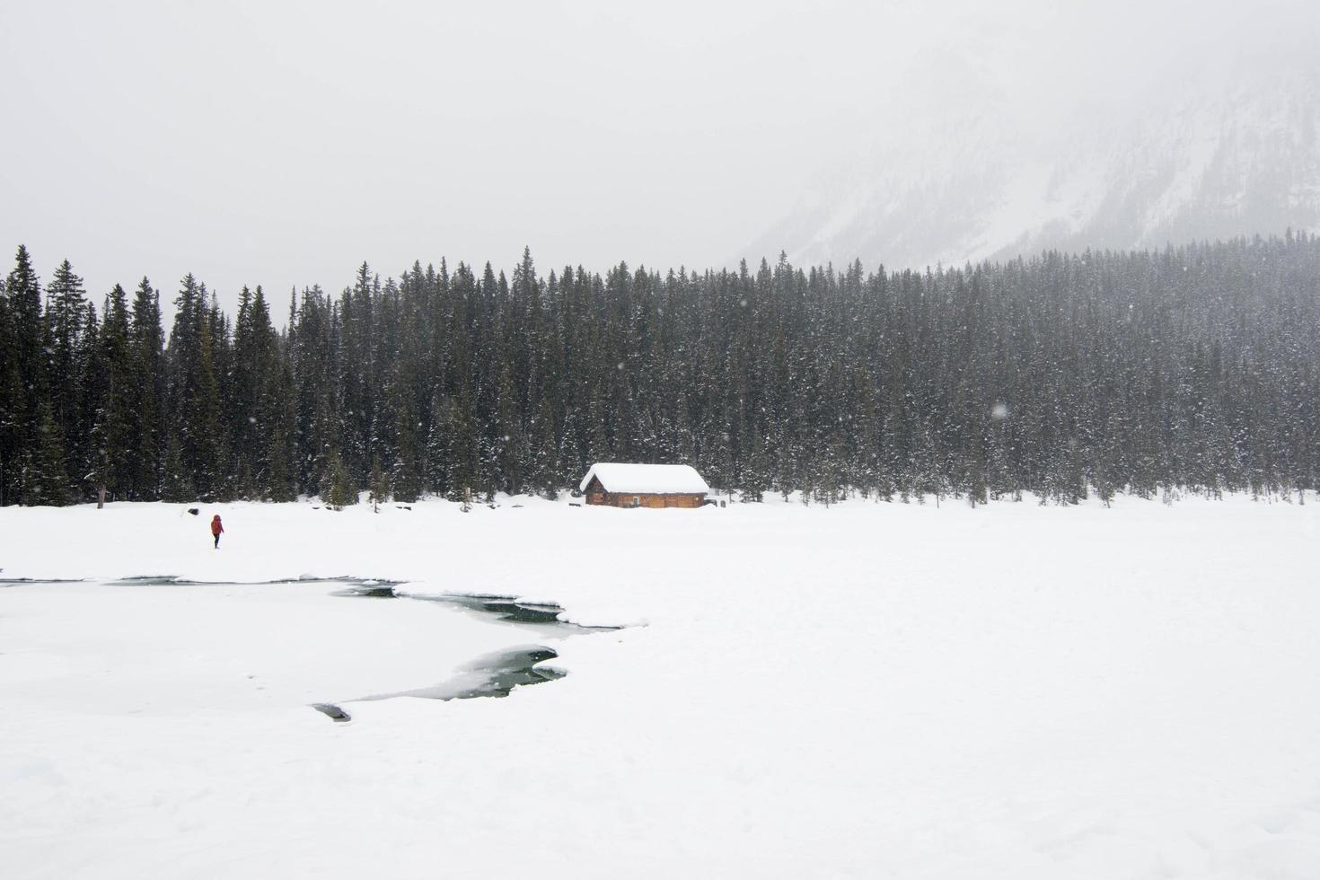 una persona vestida de rojo en un paisaje invernal. lago congelado, casa de madera y un bosque bajo la nieve. parque nacional banff, canadá foto