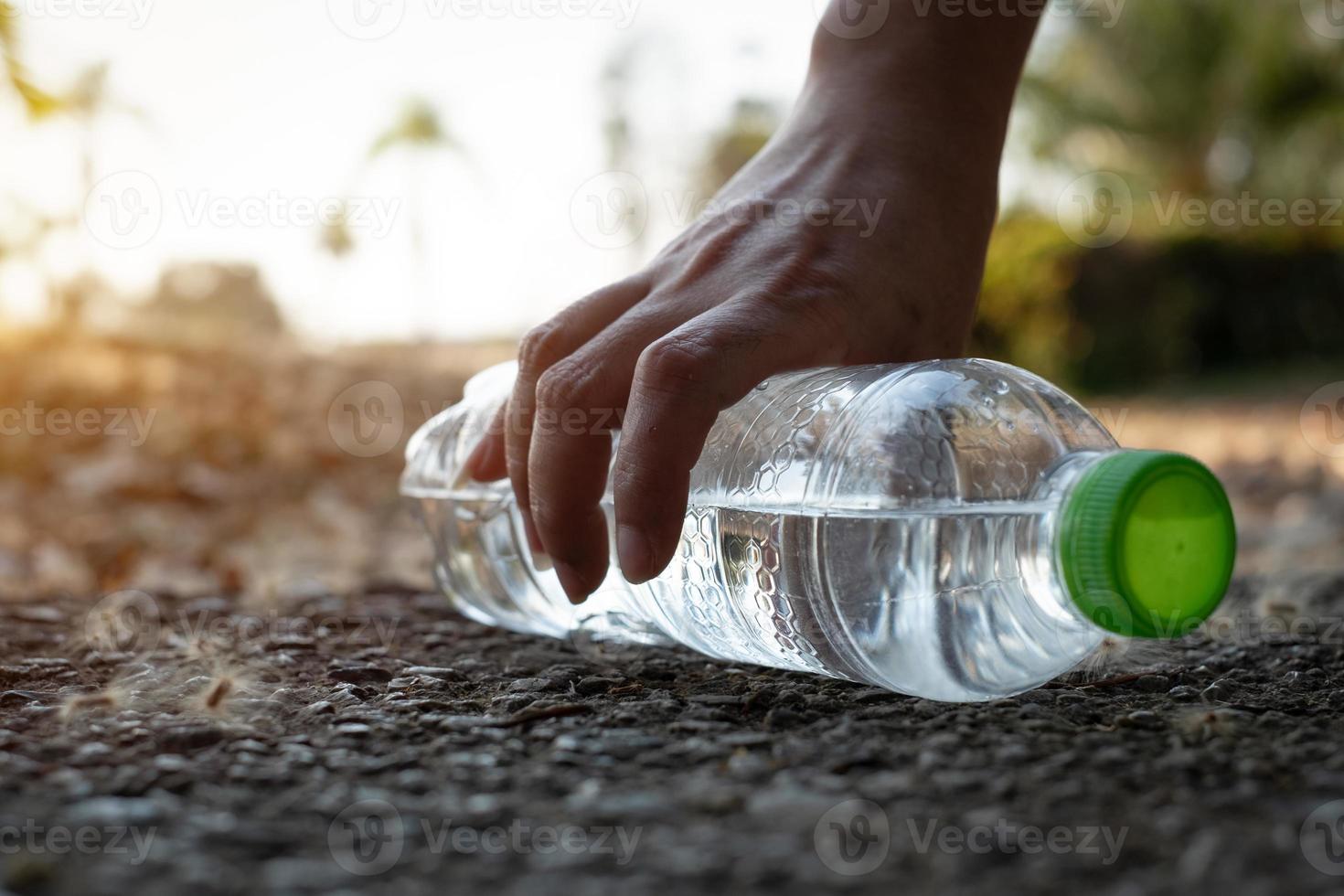 Cerrar la mano recogiendo una botella de plástico transparente, beber agua con una tapa verde en la carretera en el parque con un fondo borroso, basura que queda fuera del contenedor foto