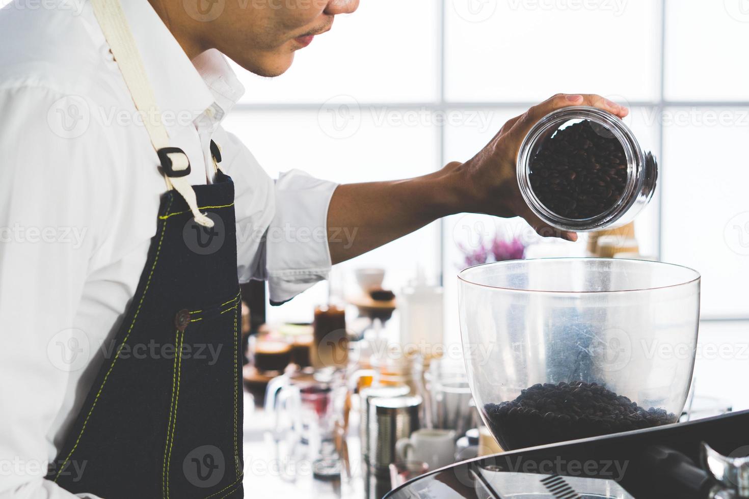 young asian man fills coffee beans into the grinder machine. barista and coffee shop concept photo