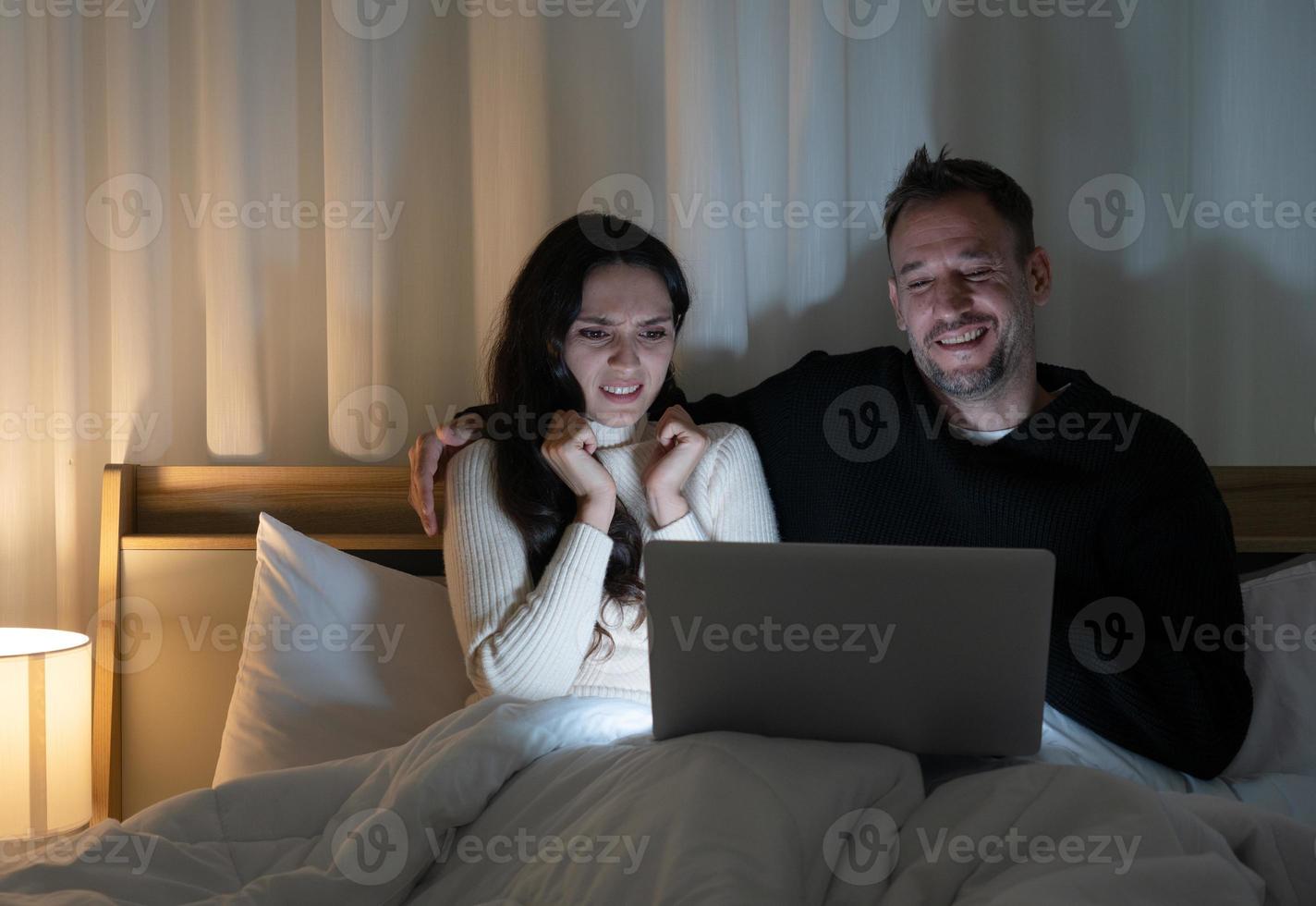 Pareja de casados viendo películas de terror del programa streming en la computadora juntos en la cama en casa por la noche foto