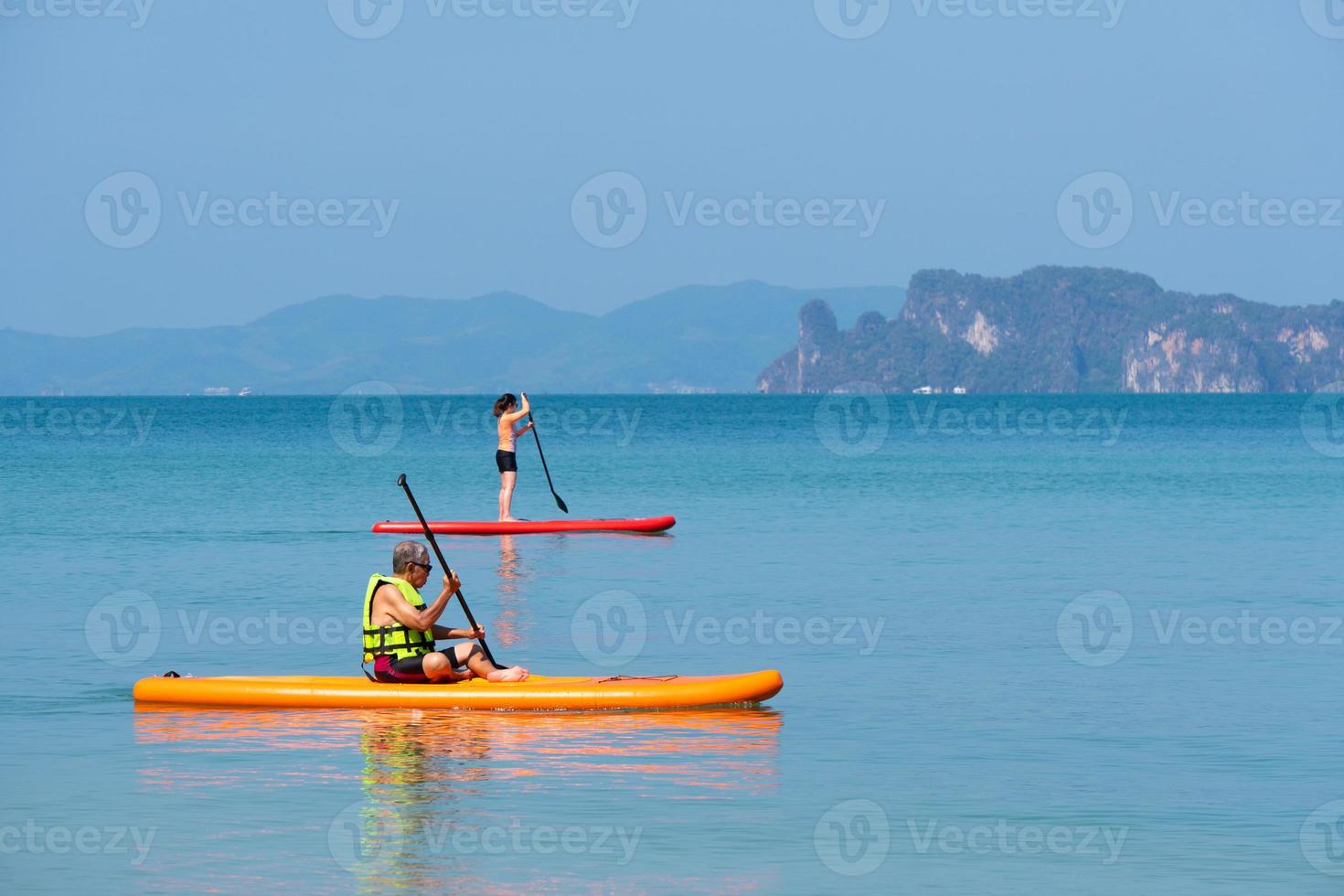 padre senior asiático jugando paddleboard de pie o sup con hija joven en el mar azul en las vacaciones de verano. concepto de familia unida foto