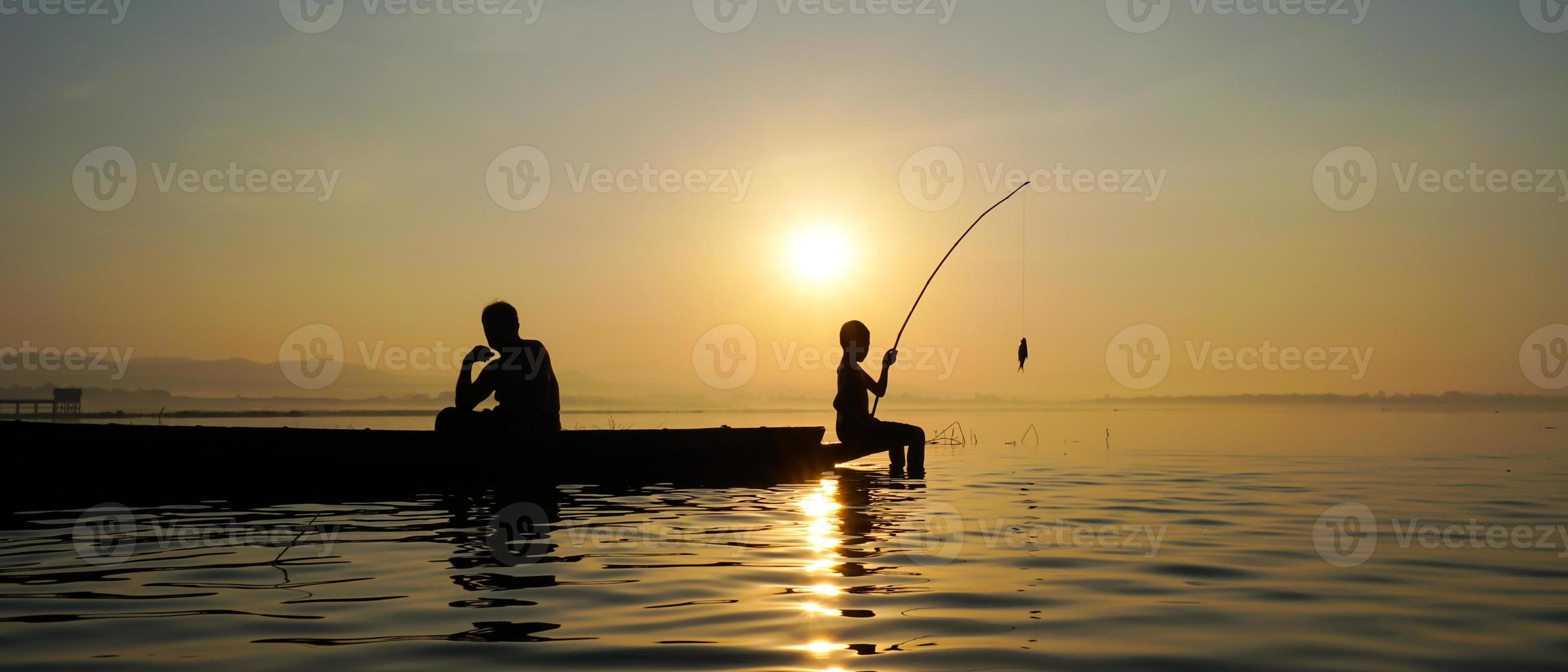 At lake side, asian fisherman sitting on boat while his son standing and  using fishing rod to catch fish at the sunrise photo