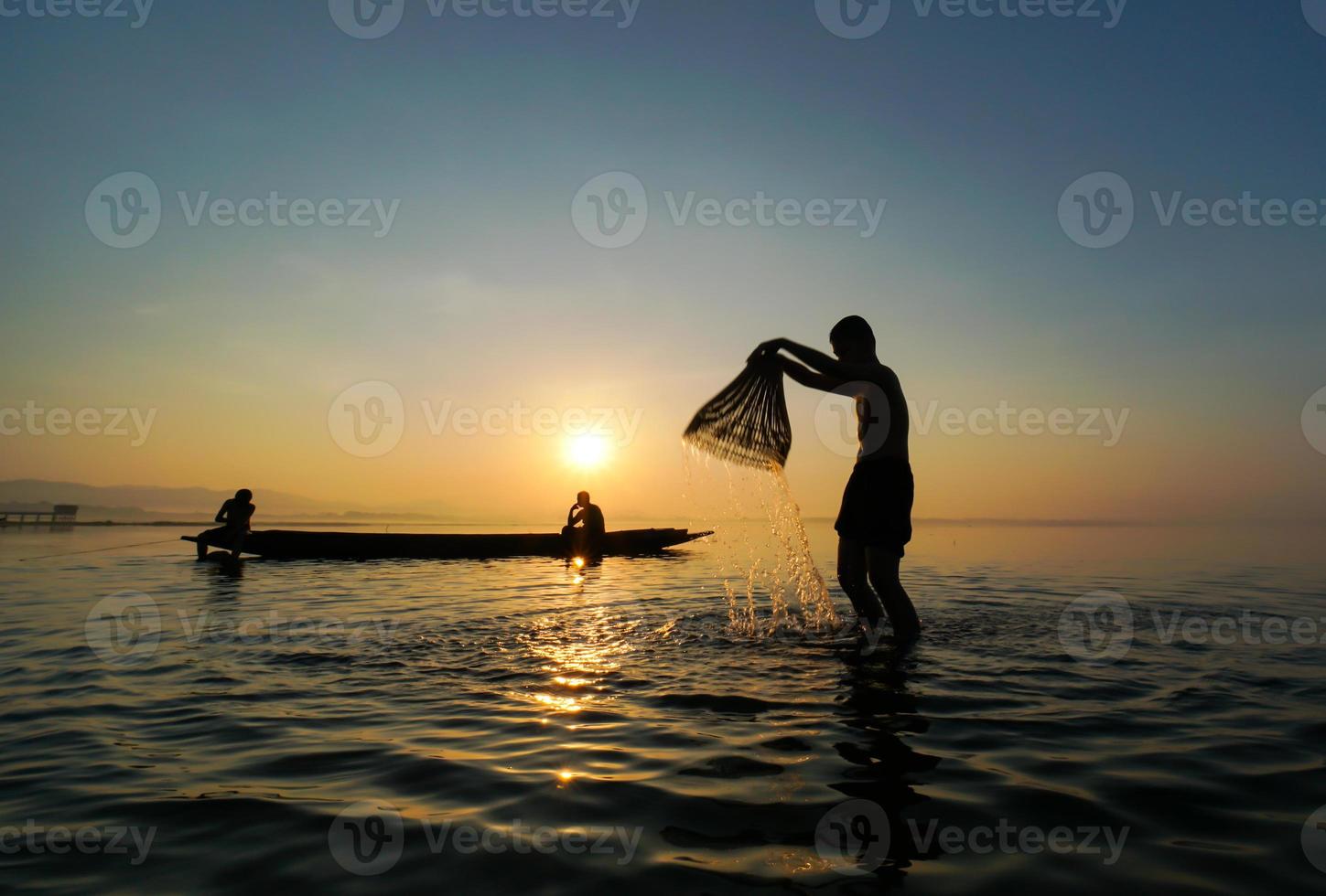 asian fisherman standing on boat and using fishing rod to catch fish while his son splashing water to each other at lake side in morning photo