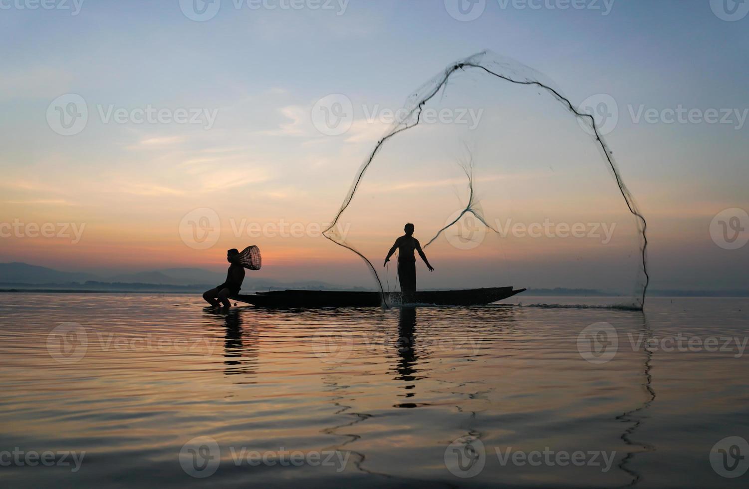 At lake side, asian fisherman sitting on boat while his son standing and  using fishing rod to catch fish at the sunrise photo