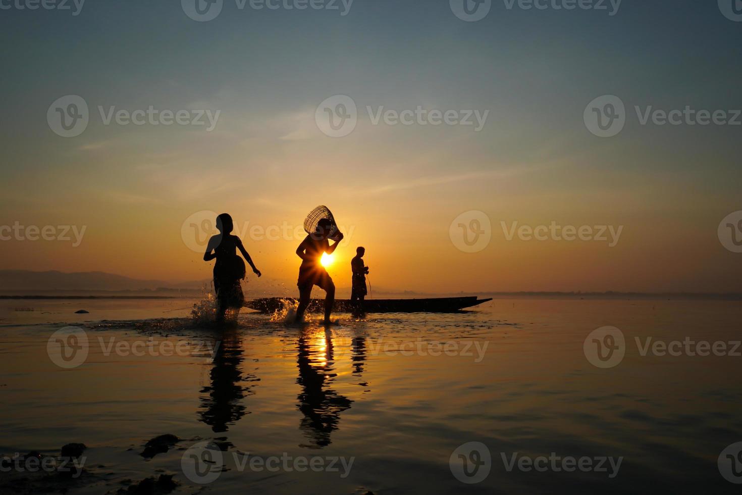 asian fisherman standing on boat and using fishing rod to catch fish while his son holding bamboo fish trap and running at lake side in morning photo