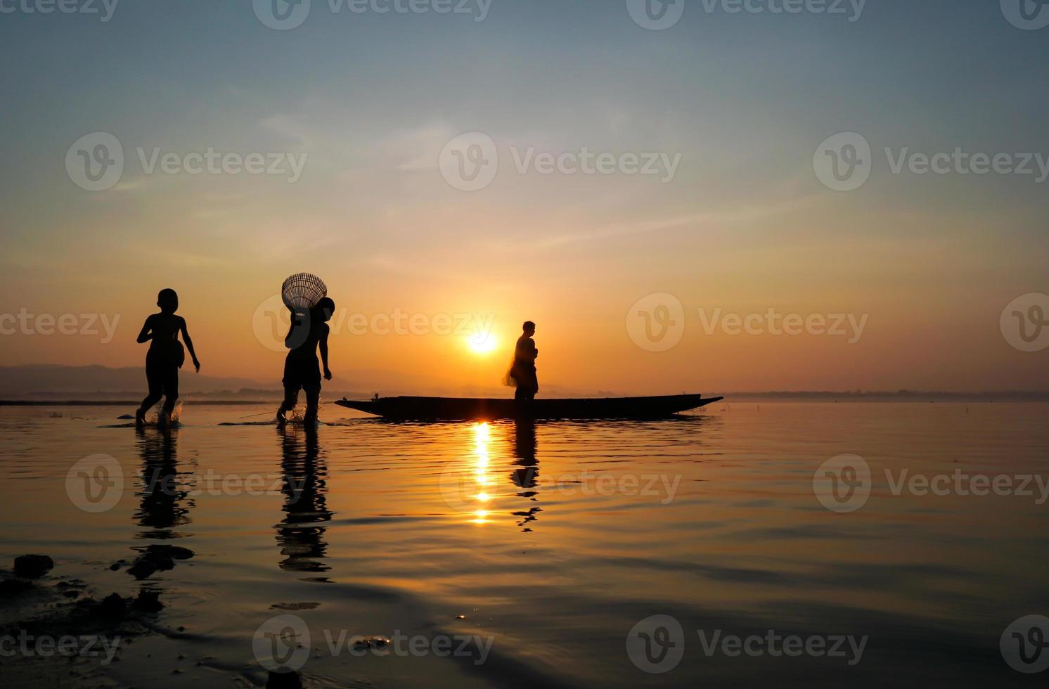 asian fisherman standing on boat and using fishing rod to catch fish while his son holding bamboo fish trap and running at lake side in morning photo