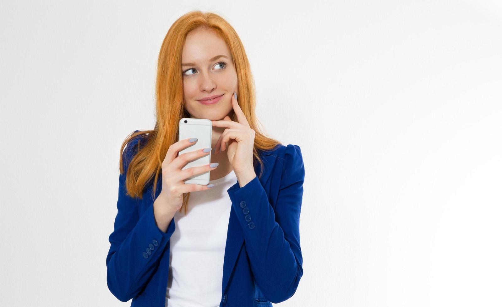 Pretty redhead girl with phone smiling isolated on white background. Young foxy female in business outfit, has amazed look, indicates with fore finger at right upper corner, shows. photo
