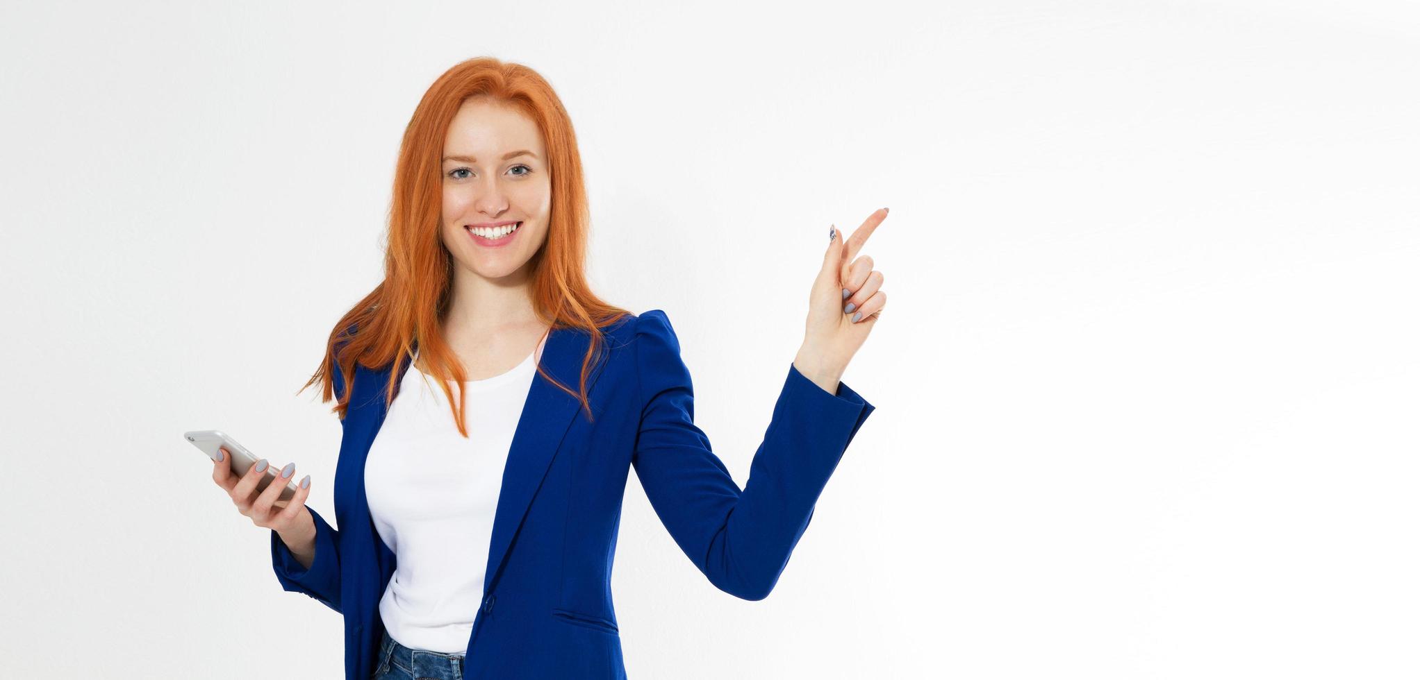 Pretty redhead girl with phone smiling pointing finger right on white background. Young foxy female in business outfit, has amazed look, indicates with fore finger at right upper corner, shows. photo