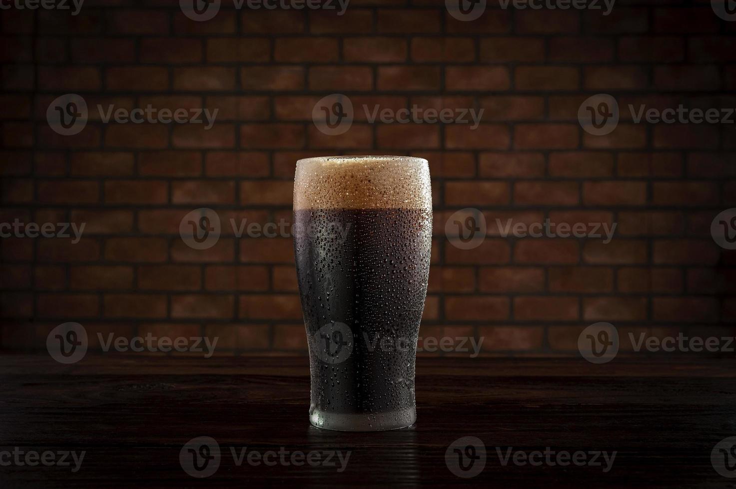 Isolated sweaty glass of refreshing dark draft beer with brick wall background. photo