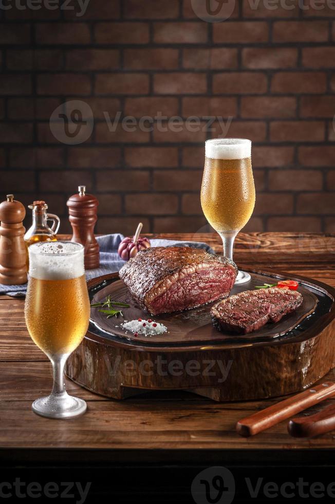 Grilled cap rump steak on wooden cutting board with two sweaty cold tulipa glasses of beer. Wooden table and bricks wall background - Brazilian picanha. photo