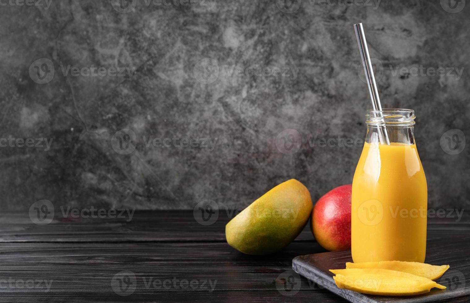 Batido de mango fresco en un vaso con una pajita de metal decorada con rodajas de mango vista frontal sobre fondo de madera oscura. foto