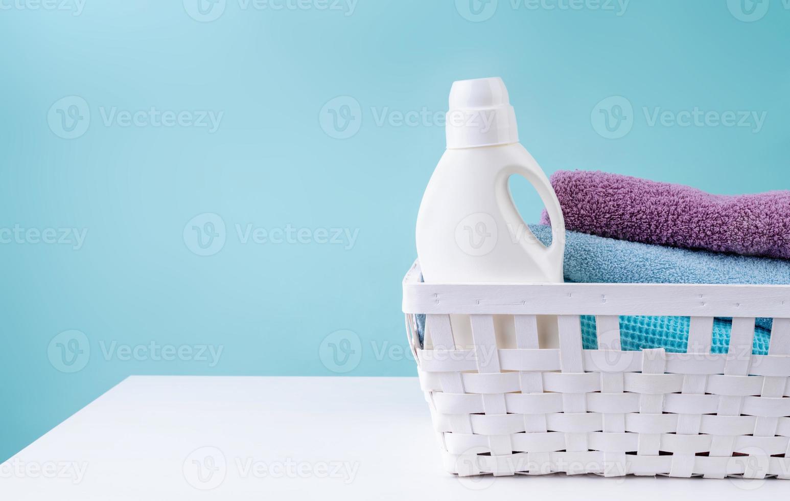 Laundry basket with a detergent bottle and a pile of clean towels on white table isolated on blue background photo