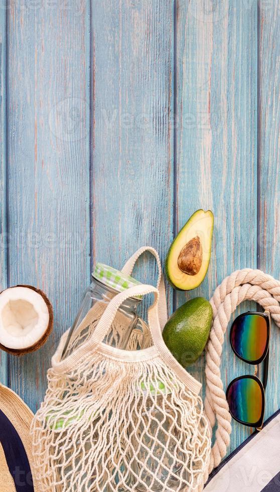 Fruta tropical en una bolsa de malla con gafas de sol y sombrero de playa, vista superior plana sobre fondo de madera azul foto