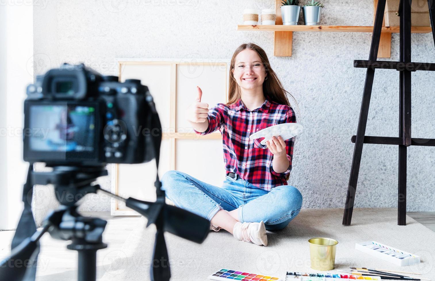 Mujer joven haciendo un video mostrando el pulgar hacia arriba para su blog sobre arte usando una cámara digital montada en un trípode foto