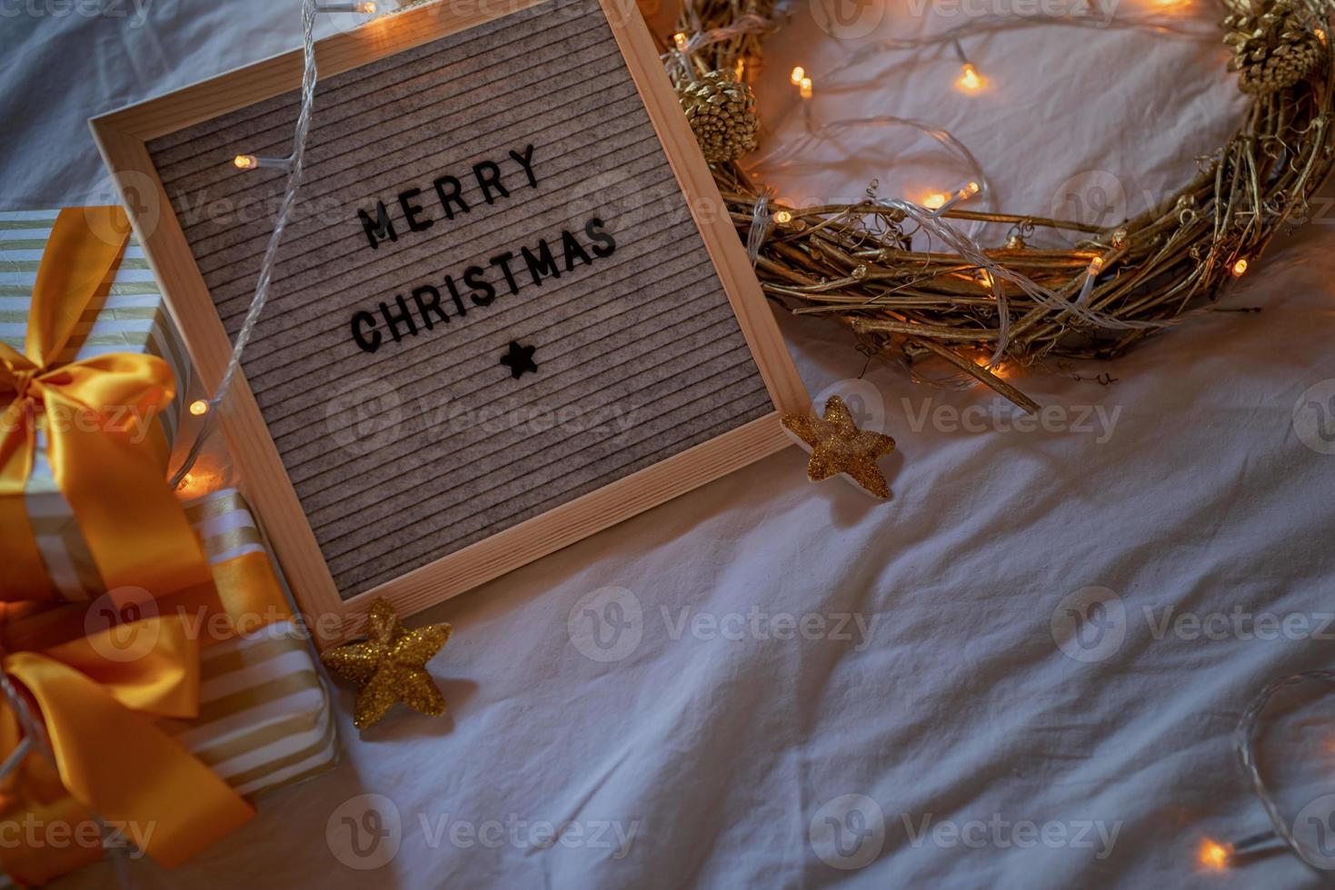 Felt letter board Merry Christmas on the bed decorated with golden wreath, lights and gift boxes photo