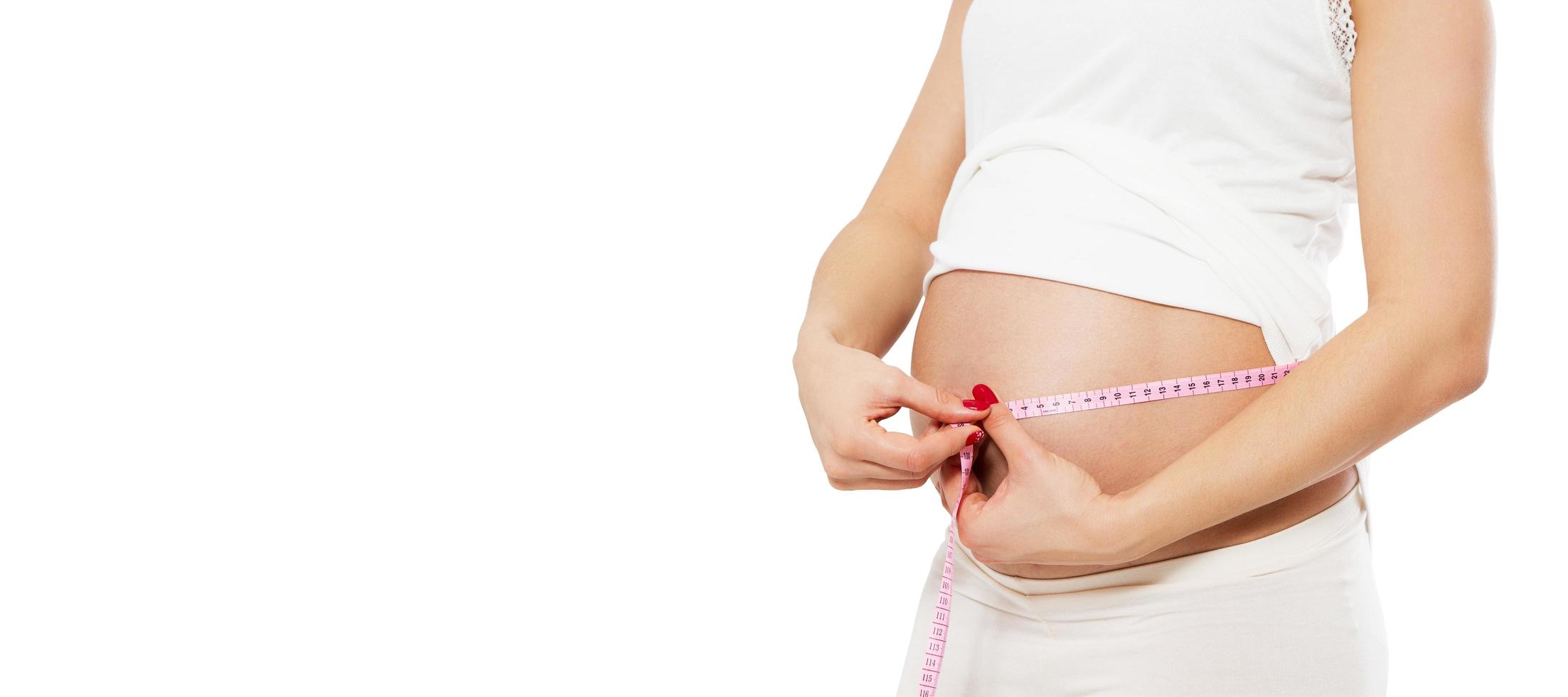 Pregnant woman measuring her belly isolated on white. Beautiful pregnant young woman in casual clothes is measuring her bare tummy cropped image photo