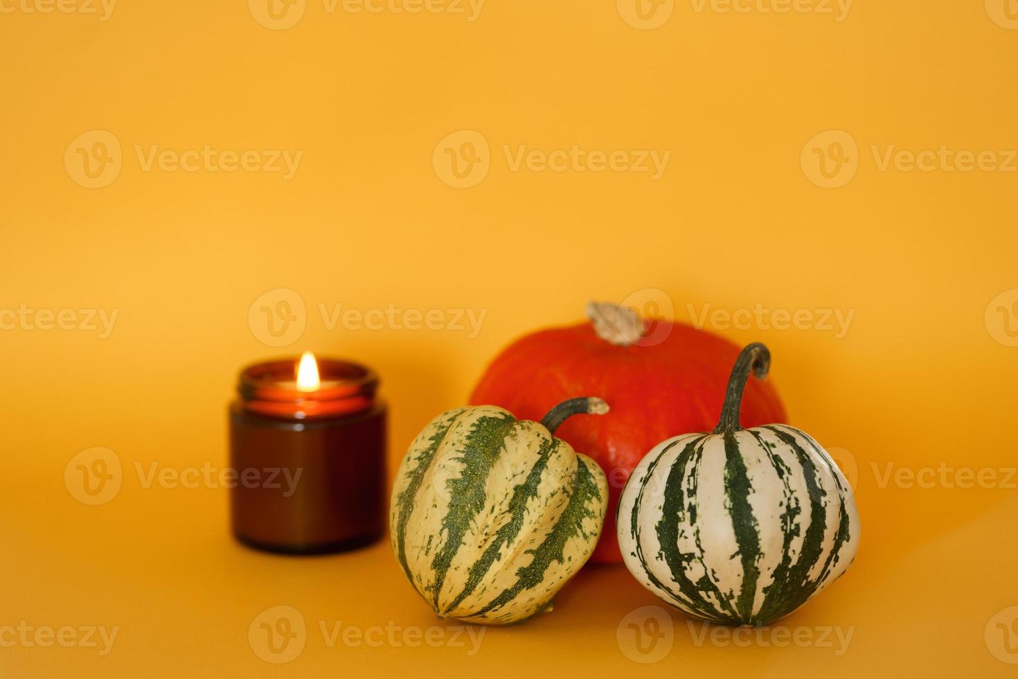 Hermosa calabaza redonda de color naranja y dos pequeñas calabazas de color blanco verdoso sobre un fondo amarillo con una vela de soja natural encendida en un frasco de vidrio oscuro. Producto organico. preparándose para halloween foto