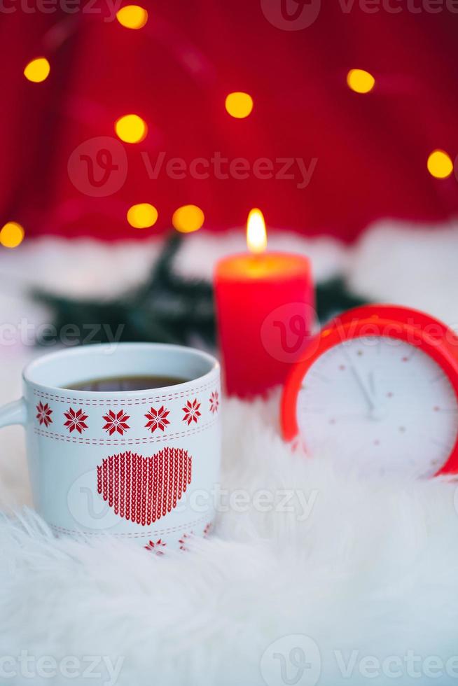 ceramic cup with red heart pattern, clock and candle with festive bokeh. happy new year. merry christmas. photo