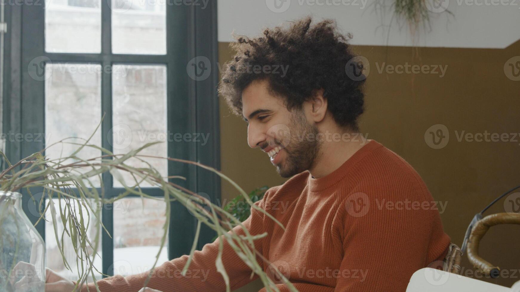 Young Middle Eastern man sits at table watching laptop in front of him, laughs photo