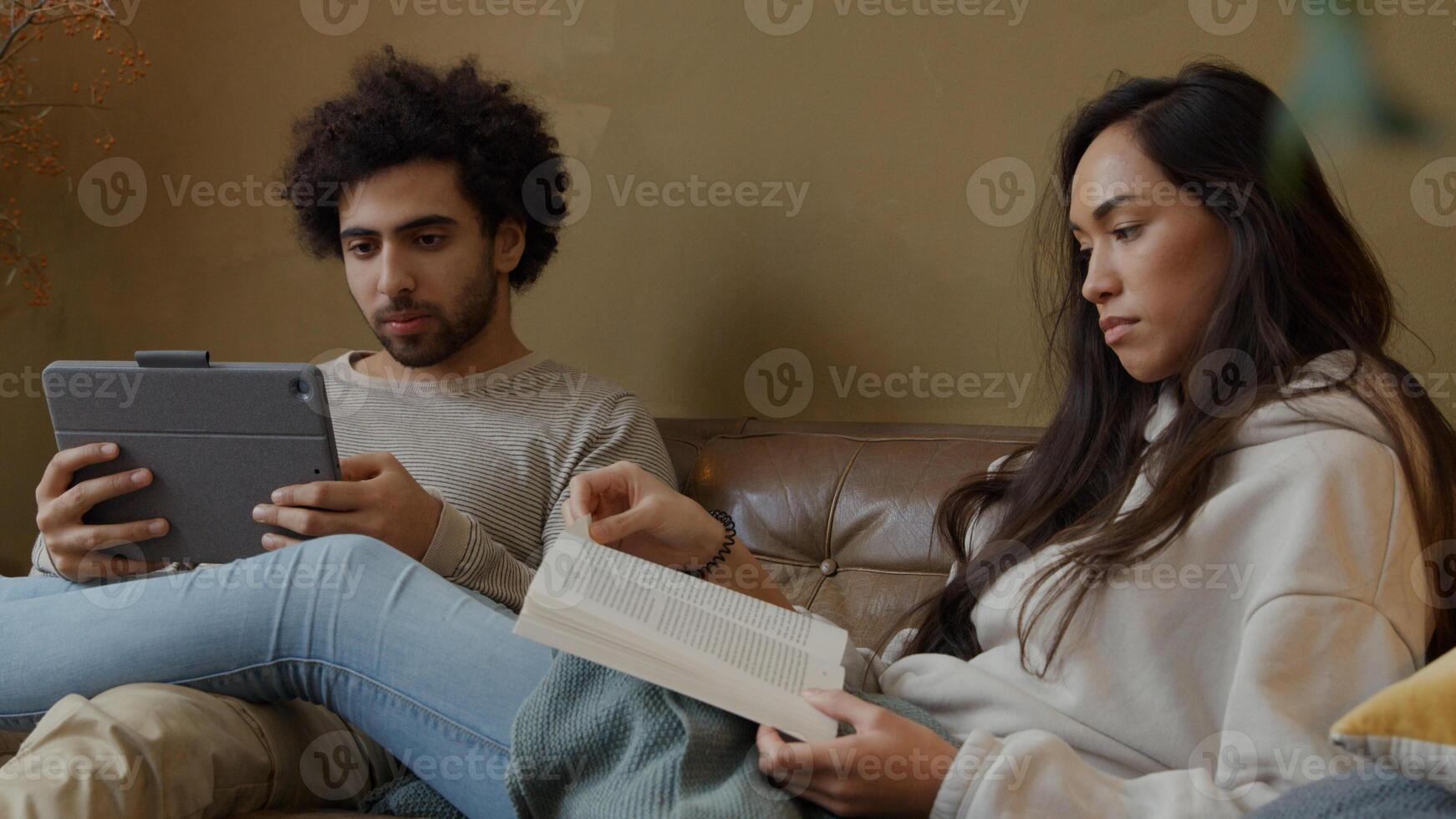 Young mixed race woman and young Middle Eastern man sitting on couch, woman reads book, man holds tablet, talking seriously photo