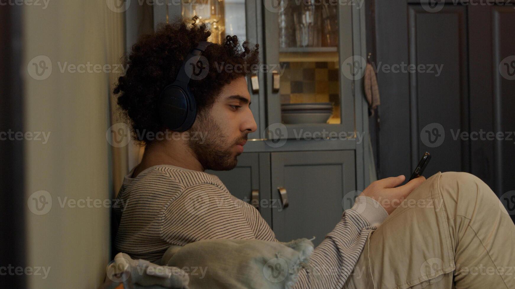 Young Middle Eastern man with headphone on his head, moves his head, sits, bent legs, holds and watches cell phone, sings a bit photo