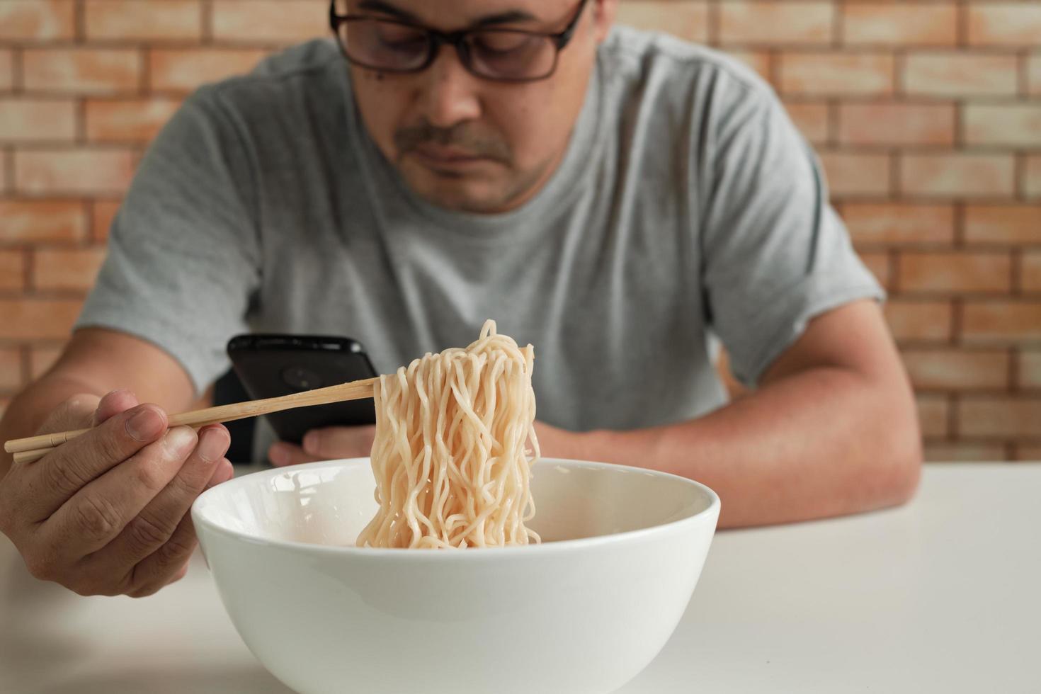 A casual Asian man on the brick wall background reading a mobile phone uses chopsticks to eat hot instant noodles meal in a white cup during lunchtime. Traditional Asian fast food cuisine lifestyle. photo