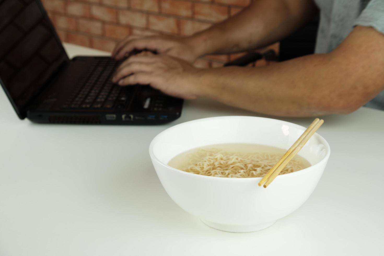 Trabajador tailandés ocupado trabajando con una computadora portátil, use palillos para comer apresuradamente fideos instantáneos durante el almuerzo de la oficina, porque es rápido, sabroso y barato. con el tiempo comida rápida asiática, estilo de vida poco saludable. foto