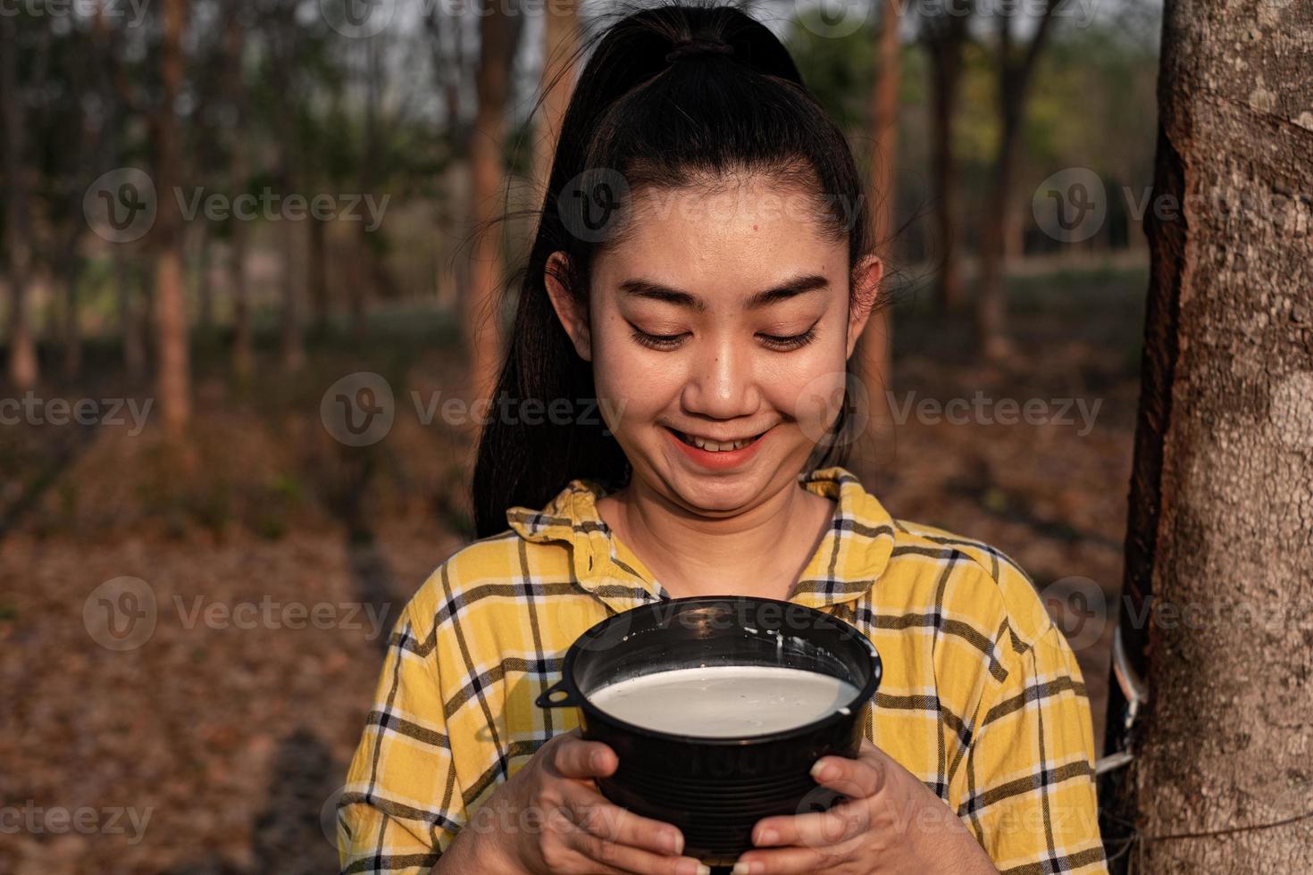 Portrait gardener young asea woman look at a full cup of raw para rubber milk of tree in plantation rubber tapping form Thailand, good farm produce photo