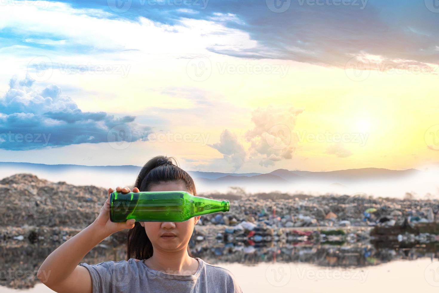 Mano de mujer sosteniendo una botella de vidrio verde para cubrir la cara en la montaña de fondo de basura grande foto