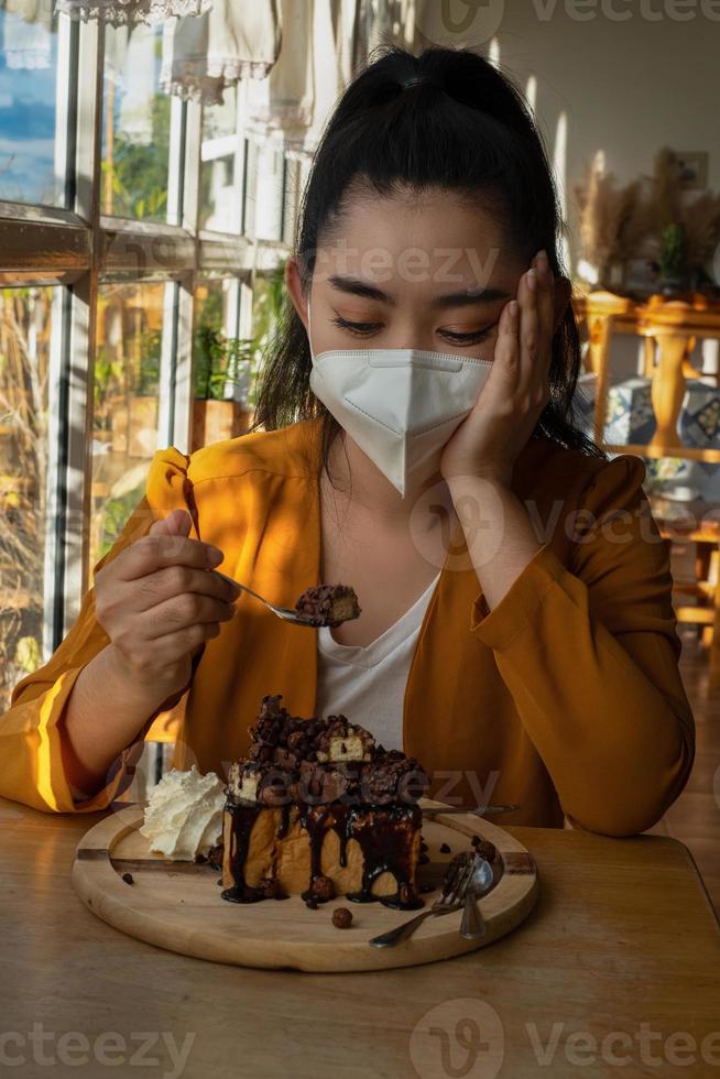 Joven mujer asiática sentada y poniéndose una mascarilla médica para protegerse de la infección por virus, enfermedades respiratorias transmitidas por el aire, miró la cuchara de pastel en la mano mientras estaba en una cafetería foto