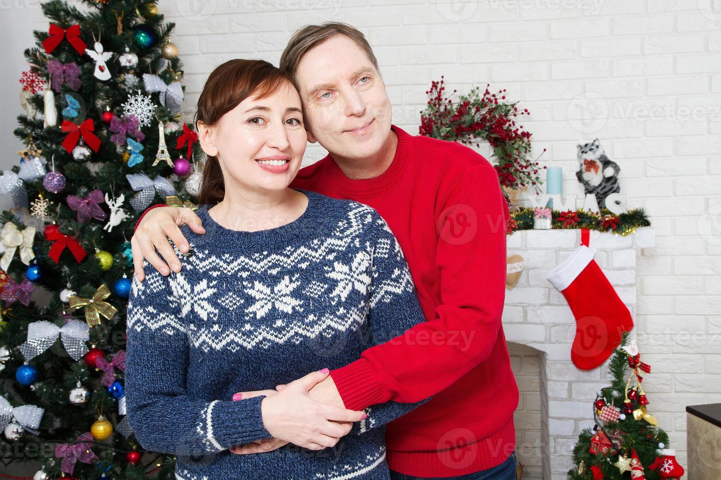 Retrato de amorosa pareja de mediana edad en la sala de estar con árbol de navidad y chimenea decorada, amorosa familia foto